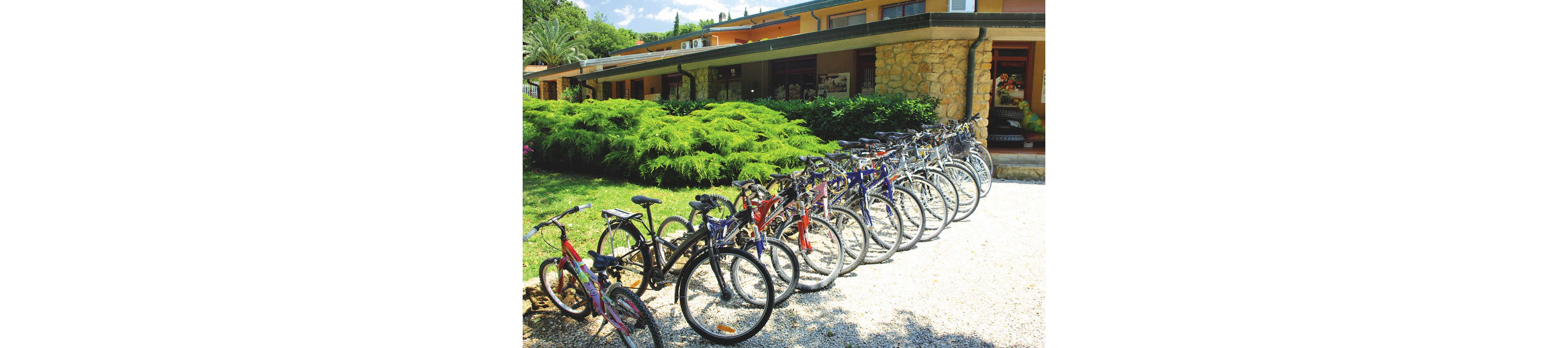 A long row of bicycles is neatly parked on a gravel area in front of a low, modern building. The structure features stone and stucco walls with a shaded walkway and large windows. Lush green shrubs and trees surround the scene, suggesting a warm, sunny day in a holiday or resort setting. The image conveys outdoor recreation and eco-friendly transportation in a relaxed environment.