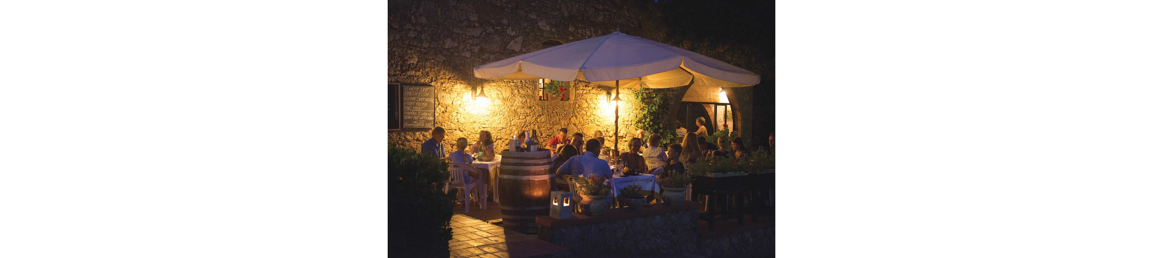 A group of people dine together at an outdoor restaurant terrace in the evening. The scene is set against a rustic stone wall with warm lantern lighting and a large white patio umbrella overhead. Diners sit at tables with white tablecloths, talking and enjoying their meals in a relaxed atmosphere. Potted plants and a wooden barrel add to the cozy, Mediterranean-style ambiance.