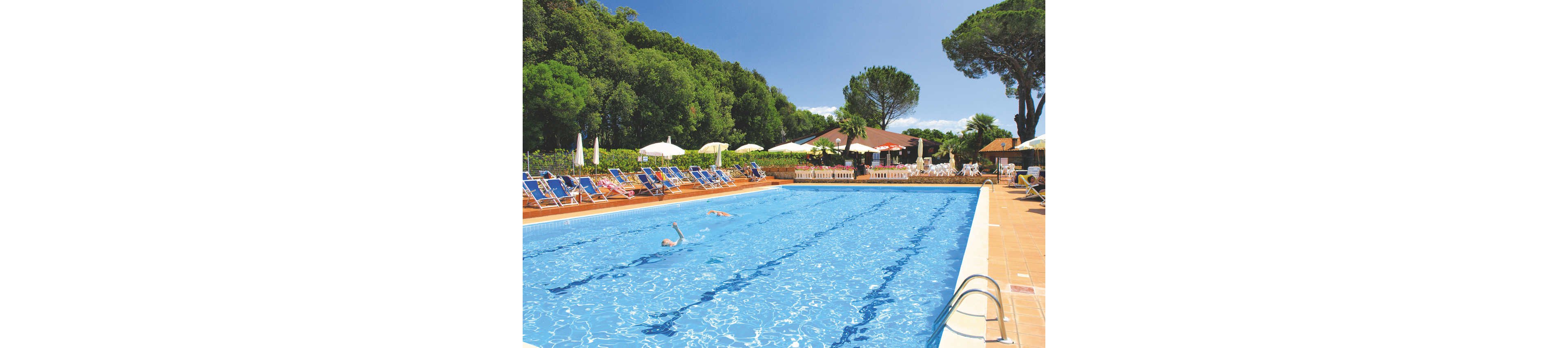 A large rectangular outdoor swimming pool stretches into the distance under a clear sky. Two people swim leisurely in the bright blue water while others relax on sun loungers along the poolside. White umbrellas, tiled decking, and a low fence frame the pool area. In the background, a building with a red roof sits beside tall green trees, creating a relaxed holiday atmosphere.