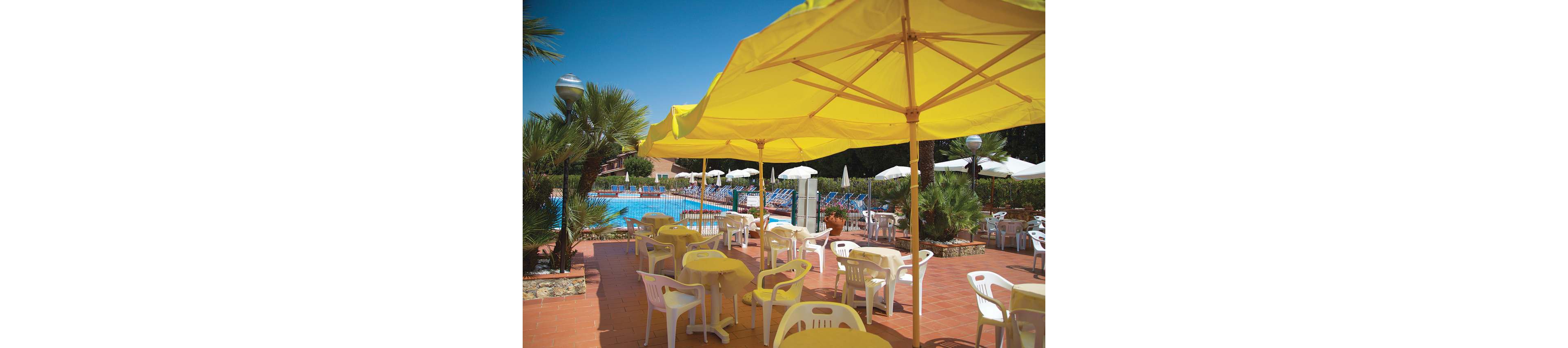 An outdoor terrace with yellow umbrellas and plastic tables overlooks a bright blue swimming pool. White lounge chairs and additional umbrellas surround the pool area in the background. Palm trees and other greenery add a tropical feel to the sunny setting. The scene suggests a relaxed vacation atmosphere at a resort or hotel.