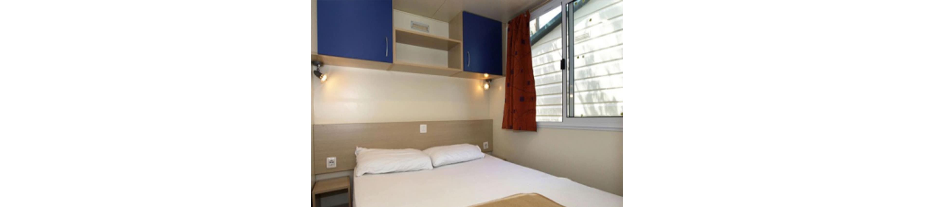 A small bedroom features a neatly made double bed with white linens against a light wood headboard. Above the bed, built-in shelving and blue cabinets provide storage and frame two wall-mounted reading lights. A large window with an orange curtain lets in natural light and shows the exterior siding of a building or mobile home. The space appears clean, minimal, and functional, suitable for a guest room or vacation rental.