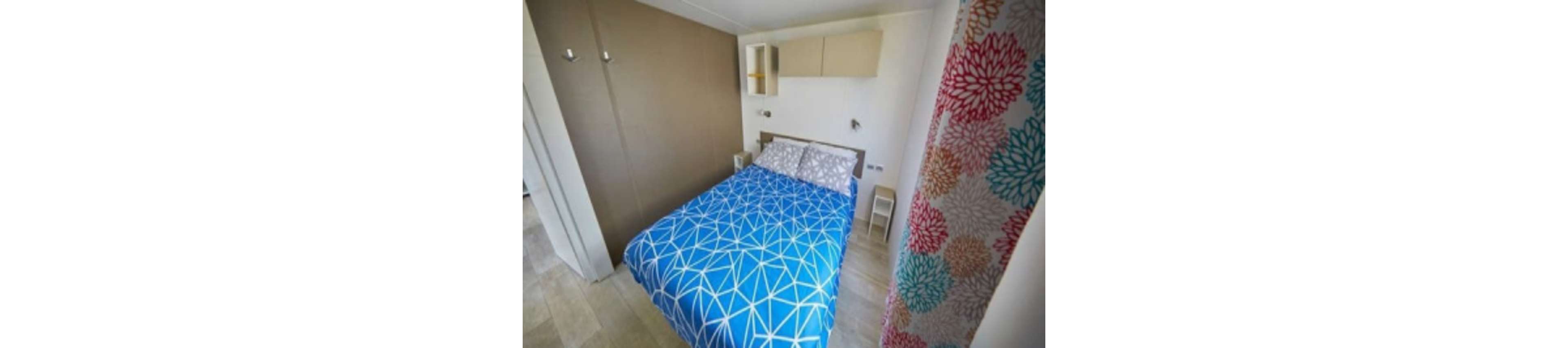 A compact bedroom features a neatly made double bed with a bright blue geometric-patterned duvet. The room has light wood flooring and a neutral accent wall behind the bed. Small white shelves and cabinets provide storage above and beside the bed. A colorful floral curtain adds contrast and warmth to the otherwise minimalist decor.