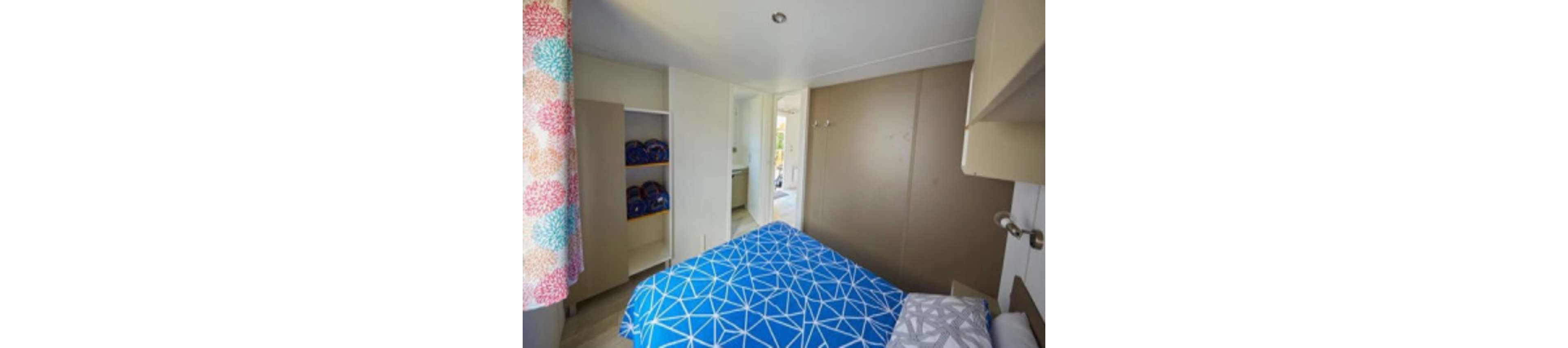 A small modern bedroom features a bed covered with bright blue geometric-patterned bedding. Built-in shelves with neatly stacked items sit to the left, and a doorway opens to an adjacent room or hallway. Neutral walls and white cabinetry create a clean, minimalist look. Colorful patterned curtains add a touch of warmth and contrast to the otherwise simple space.
