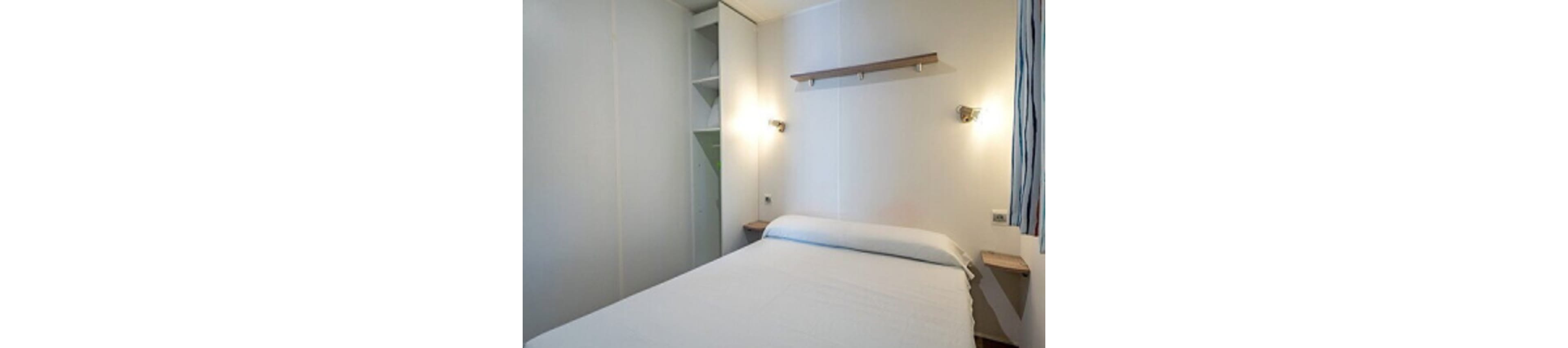 A compact bedroom features a neatly made bed with a white bedspread against a plain wall. Built-in white wardrobes with open shelving occupy one side of the room. Two wall-mounted lights provide warm illumination, and a simple wooden shelf hangs above the bed. A narrow window with striped curtains adds a touch of color to the otherwise minimalist space.