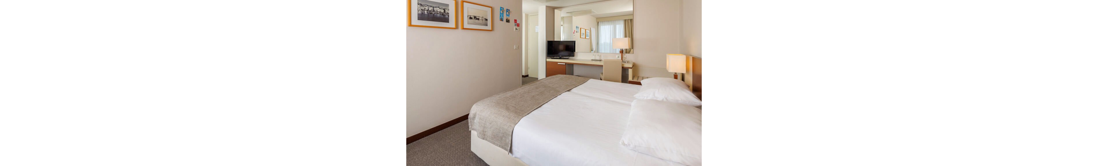 A neatly made double bed with white linens and a beige throw dominates a modern hotel room. In the background, a flat-screen television sits on a desk with a chair, creating a small workspace. Soft lighting from bedside lamps and a large window with curtains give the space a bright, welcoming feel. Framed artwork on the walls and neutral tones contribute to the clean, contemporary style.