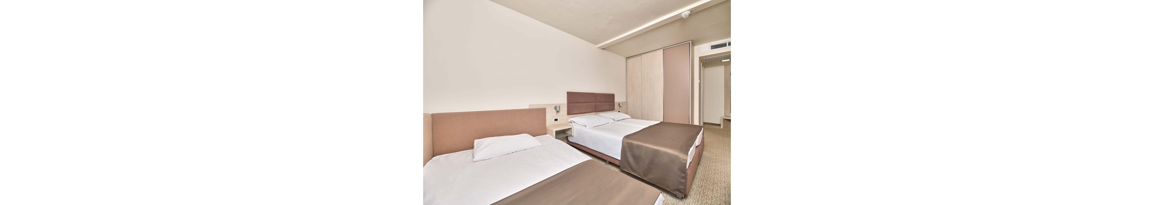 A contemporary hotel room features two neatly made beds with brown bedspreads and white linens. The space is bright and minimal, with light-colored walls and built-in wardrobe doors in neutral tones. Wall-mounted reading lights and a small bedside shelf sit between the beds, adding functional comfort. The overall design is clean and simple, creating a calm and inviting atmosphere for guests.