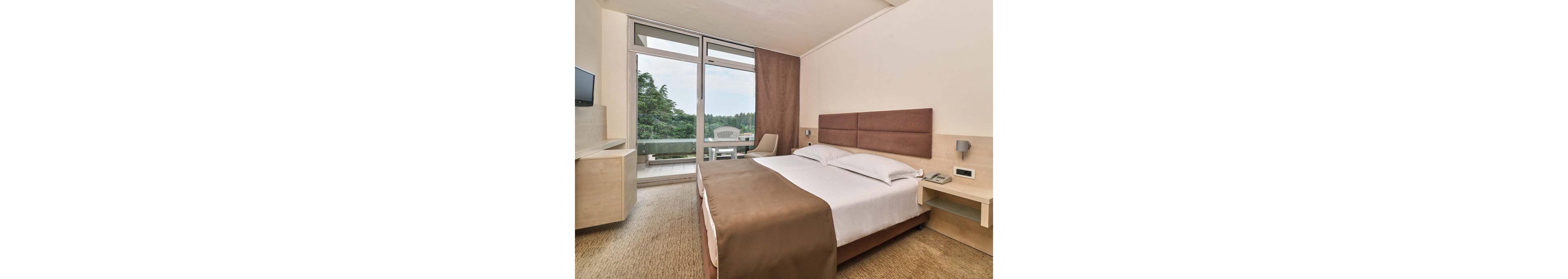 A contemporary hotel room features a large double bed with brown and white bedding. The space includes light wood furniture, a wall-mounted television, and bedside tables with lamps and a telephone. Floor-to-ceiling glass doors open onto a balcony with outdoor chairs and a view of green trees. Neutral tones and simple decor create a clean, comfortable atmosphere suitable for travelers.
