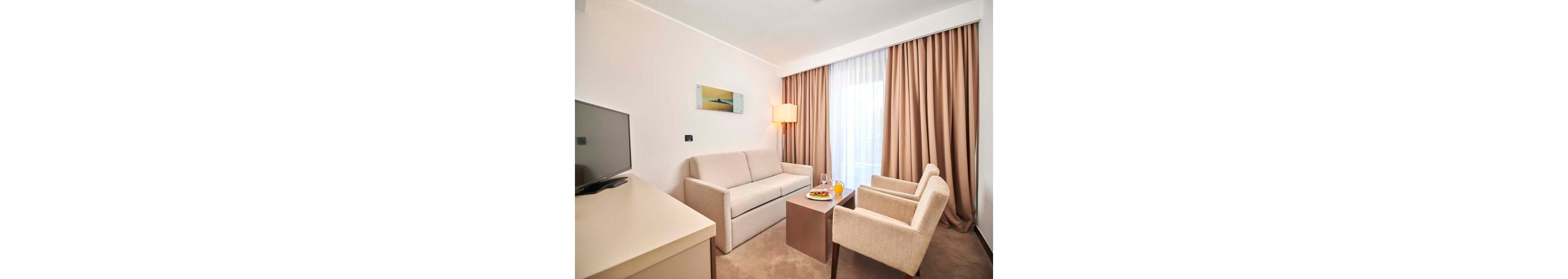 A compact living room features a beige sofa and two matching armchairs around a small wooden coffee table. The space is brightly lit by a floor lamp and natural light coming through a large window with sheer white curtains and heavier beige drapes. A flat-screen TV sits on a light-colored desk opposite the seating area, and a small wall art piece hangs above the sofa. On the coffee table, a plate of food and drinks in glasses add a touch of hospitality and comfort.