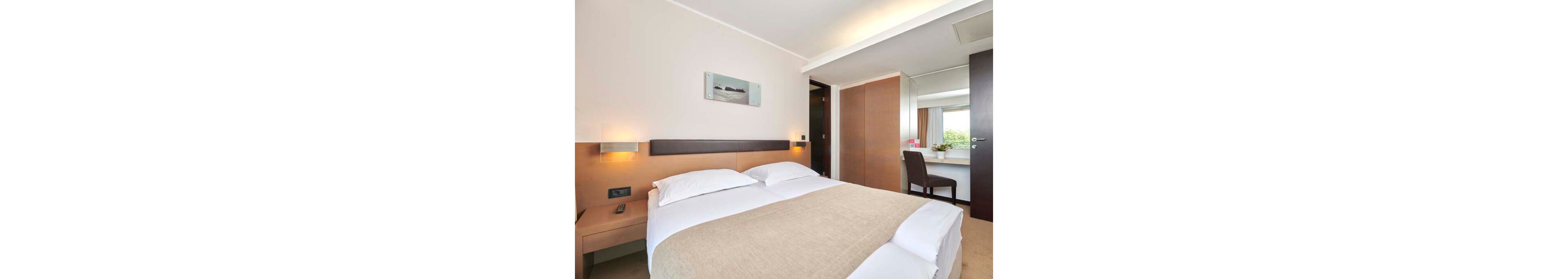 A contemporary hotel room features a neatly made double bed with white linens and a beige bedspread. Warm wall-mounted lights illuminate the headboard area and a minimalist artwork above the bed. To the right, a desk with a chair sits near a window with curtains, providing a small workspace. The overall design is clean and modern, with neutral tones and simple furnishings creating a calm, inviting atmosphere.