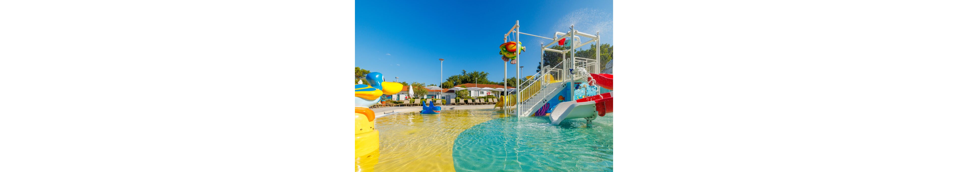 A vibrant children’s water play area features shallow pools and colorful slides under a clear blue sky. Animal-shaped water toys and a multi-level play structure with stairs and sprinklers create a fun, family-friendly setting. Small houses and trees in the background suggest a holiday resort or campground environment. Bright yellow and turquoise water surfaces enhance the playful, sunny atmosphere.