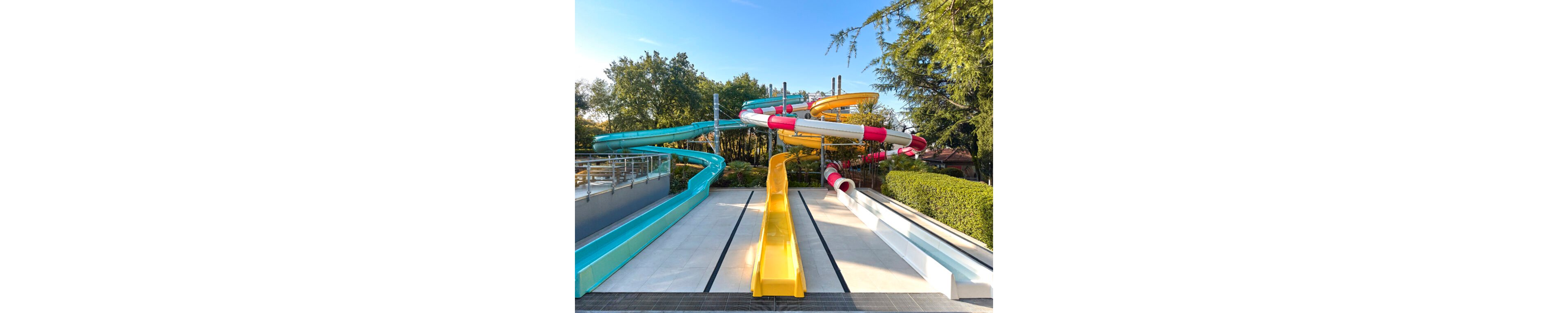 A set of bright water slides is shown in an outdoor water park, with multiple chutes in yellow, blue, white, and red. The slides curve and twist above the ground before ending in parallel lanes on a tiled platform. Surrounding trees and greenery frame the scene under a clear blue sky, suggesting a sunny day. The area appears clean and empty, ready for visitors.