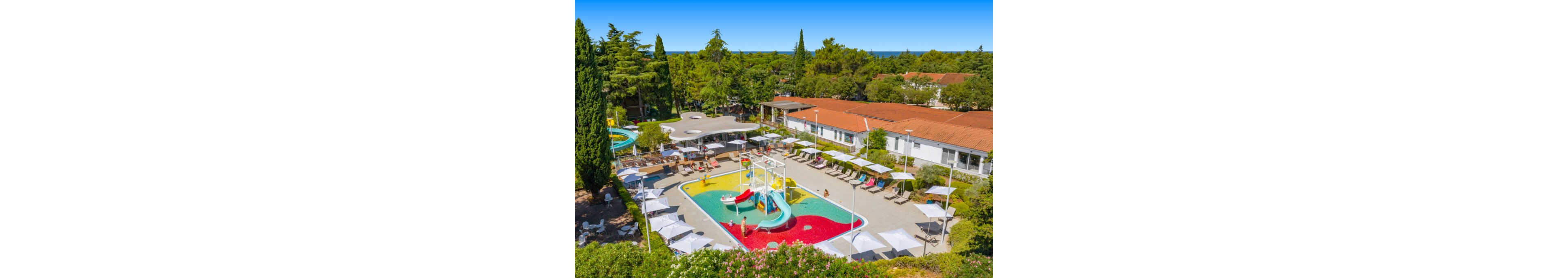 A vibrant outdoor water playground is set within a holiday resort complex surrounded by trees. Shallow pools with slides, sprayers, and play structures create a fun area for children and families. Sun loungers and white parasols line the paved deck around the water features, offering space to relax. Low white buildings with red-tiled roofs frame the scene under a clear blue sky.