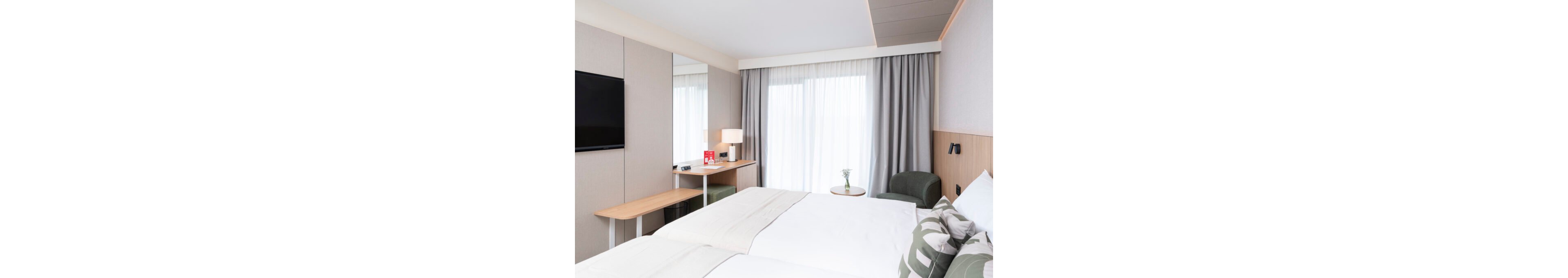A contemporary hotel room features a neatly made double bed with light bedding and patterned pillows. A wall-mounted flat-screen TV faces the bed, and a minimalist wooden desk with a lamp sits beside the window. Floor-to-ceiling curtains cover a large glass door or window, allowing soft natural light to fill the space. A small round side table with a single flower and a green armchair add a cozy touch to the clean, neutral interior.