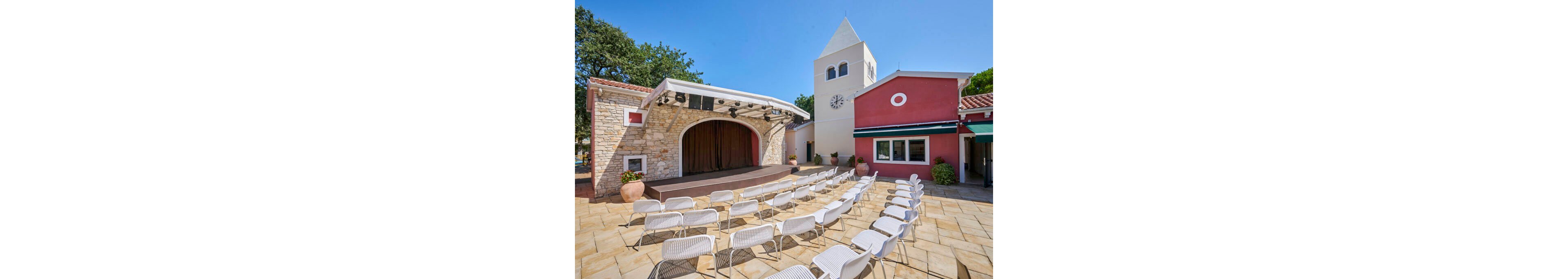 An outdoor courtyard features a small performance stage framed by stone walls and a curved arch. Rows of white chairs are neatly arranged facing the stage, suggesting a venue ready for a show or presentation. A tall tower with a clock and adjacent colorful buildings create a Mediterranean village atmosphere. The scene is brightly lit under a clear blue sky, with potted plants adding a touch of greenery.