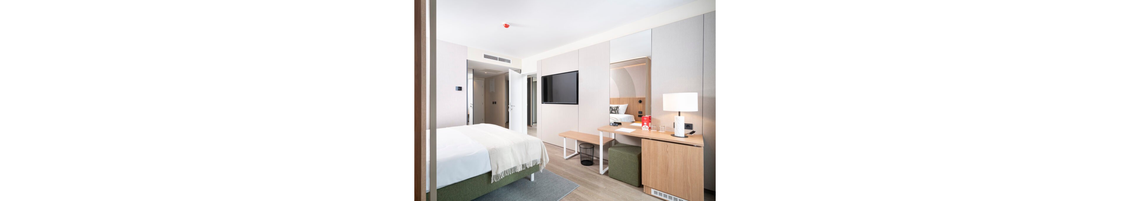 Contemporary hotel room interior featuring a neatly made double bed with a light throw blanket. A wall-mounted flat-screen TV faces the bed, and built-in wardrobes line the wall. A compact wooden desk with a lamp, chair, and small ottoman creates a functional workspace. The room is decorated in neutral tones with soft lighting, giving a clean and minimalist feel.
