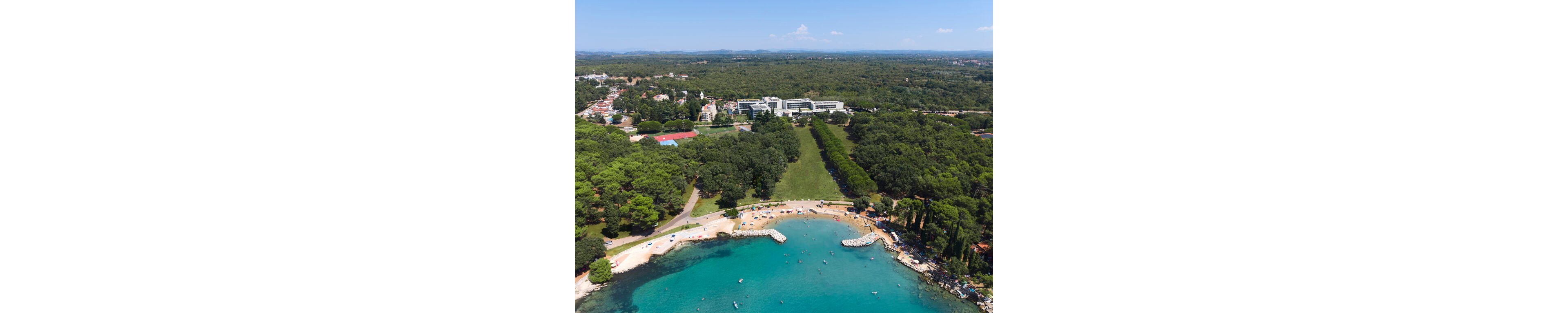 A wide aerial view shows a coastal resort with a sandy beach and turquoise water in the foreground. Sunbathers and umbrellas line the curved shoreline, with small boats visible in the calm bay. Behind the beach, dense green forest and lawns lead up to modern white buildings and a town area. The scene extends to distant hills under a clear blue sky, suggesting a sunny vacation destination.