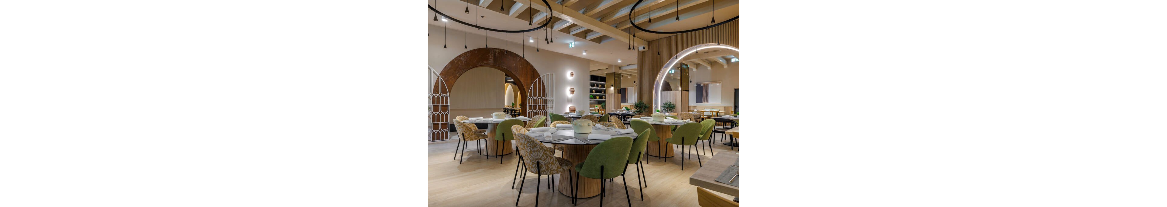 Contemporary restaurant dining room featuring round tables set with white napkins and tableware. Green and patterned upholstered chairs surround the tables, creating a stylish and inviting seating area. Architectural arches, wood paneling, and decorative lighting add visual interest to the spacious interior. Soft, warm illumination and neutral tones contribute to a relaxed yet sophisticated atmosphere.