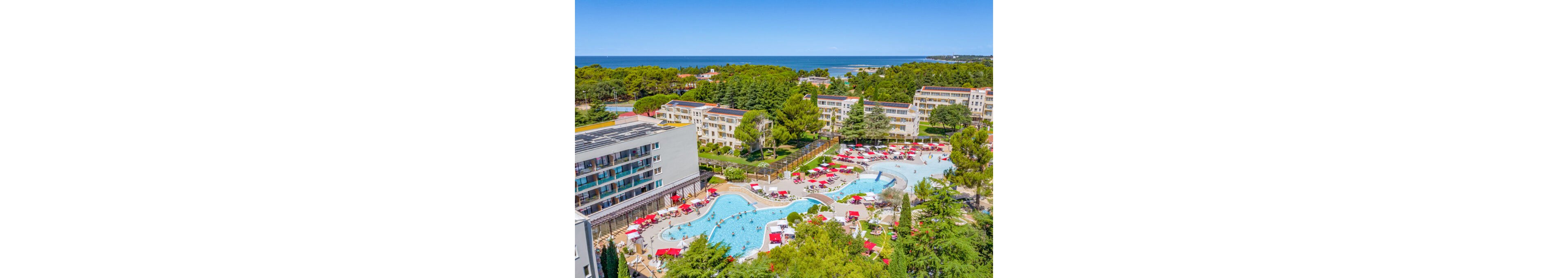 A large seaside resort complex features multiple outdoor swimming pools surrounded by sun loungers and umbrellas. Modern multi-story buildings and low-rise apartments are nestled among lush green trees and landscaped gardens. Guests relax around the curved pools, creating a lively vacation atmosphere under clear blue skies. In the distance, the coastline and calm sea frame the resort, emphasizing its coastal holiday setting.