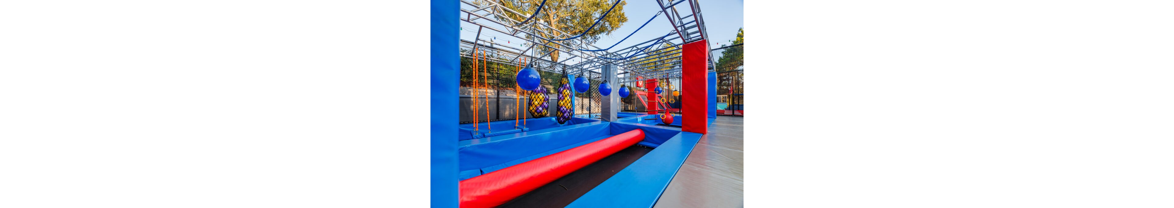 Image of a brightly colored outdoor obstacle course inspired by ninja training. The structure features hanging balls, rope elements, padded beams, and netted obstacles within a metal frame. Blue and red safety padding covers the floor and sides, with trees and fencing visible in the background. The scene is empty of people, emphasizing the equipment and playful, energetic environment.