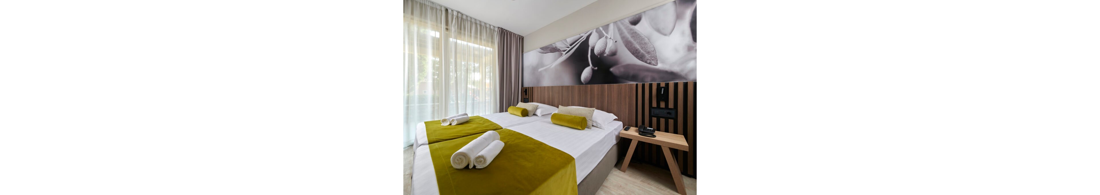 Contemporary hotel room interior featuring two neatly made twin beds with white linens and olive green runners. Rolled white towels are placed at the foot of each bed, complemented by matching cylindrical cushions. A large black-and-white wall mural with botanical imagery decorates the headboard area, adding visual interest. Floor-to-ceiling curtains filter natural light from the window, while a wooden bedside table holds a telephone and remote control, enhancing the functional, welcoming atmosphere.