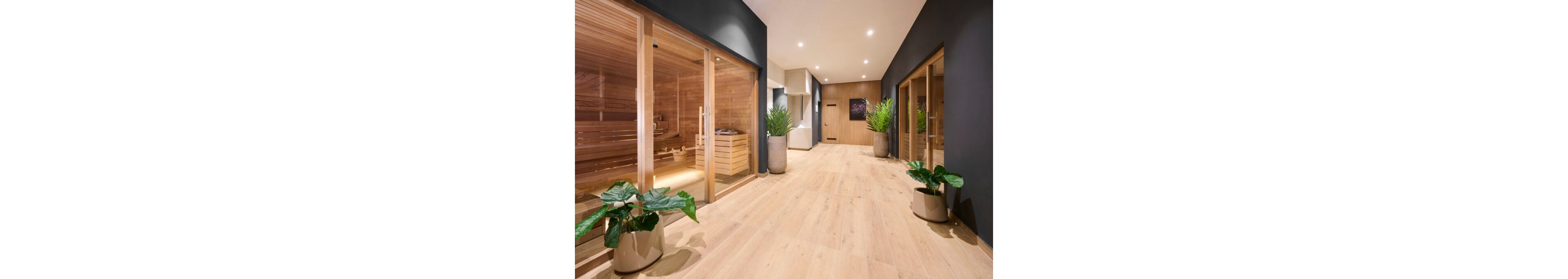 A contemporary spa hallway features multiple wooden sauna rooms with glass doors along one side. The space is finished with light wood flooring and dark accent walls, creating a clean, minimalist look. Large potted green plants add natural contrast and soften the modern interior. Recessed ceiling lights evenly illuminate the corridor, emphasizing the warm, inviting atmosphere.
