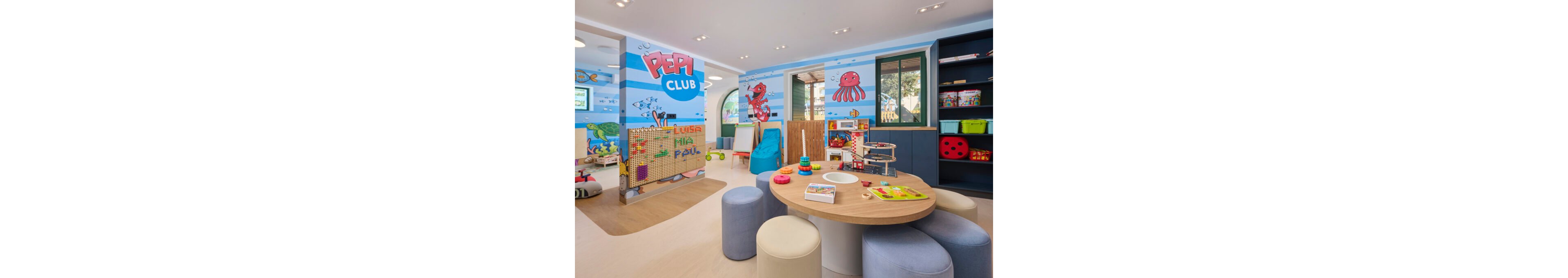 Brightly decorated Pepi Club children’s playroom features round tables, soft stools, and various toys. The walls are painted with underwater-themed illustrations including fish, an octopus, and the Pepi Club name. Activity stations with building blocks, pegboards, and art supplies invite creative play. Large windows and open shelving filled with games and books create a welcoming, organized space for kids.
