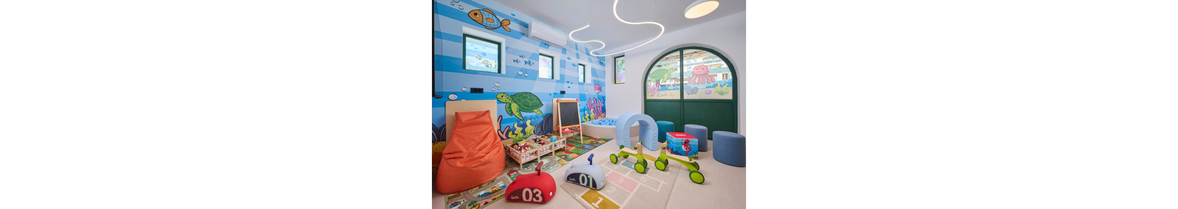 Bright indoor playroom decorated with an ocean-themed wall mural featuring fish, turtles, and sea plants. The room includes soft play toys, a hopscotch rug, beanbag seating, and a small activity table with blocks. Large arched doors and small windows let in natural light, enhancing the playful atmosphere. Visible numbers on toys and floor include 01, 02, 03, and 1, 2, 3.