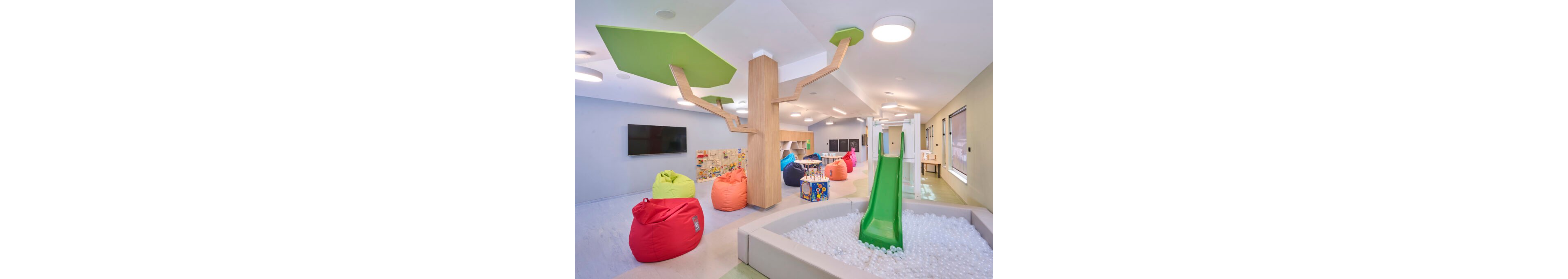 Bright contemporary children’s playroom featuring a green slide into a white ball pit. Colorful bean bags and soft seating are arranged around the room, creating a relaxed area for kids to sit and play. A wooden column with stylized green tree panels adds a playful decorative element to the ceiling. Shelves with toys and books line the walls, and large windows and ceiling lights provide ample natural and artificial light.