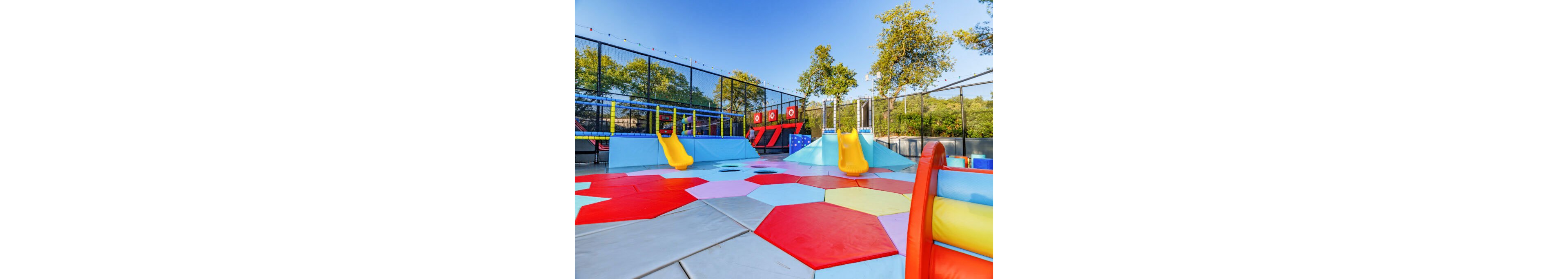 A bright outdoor playground features multiple yellow slides and soft geometric floor mats. The play area includes hexagonal and polygonal shapes in red, pink, blue, and gray, creating a vibrant pattern. Net fencing surrounds the space, with trees and greenery visible in the background under a clear blue sky. The scene appears empty, emphasizing the equipment and colorful design.