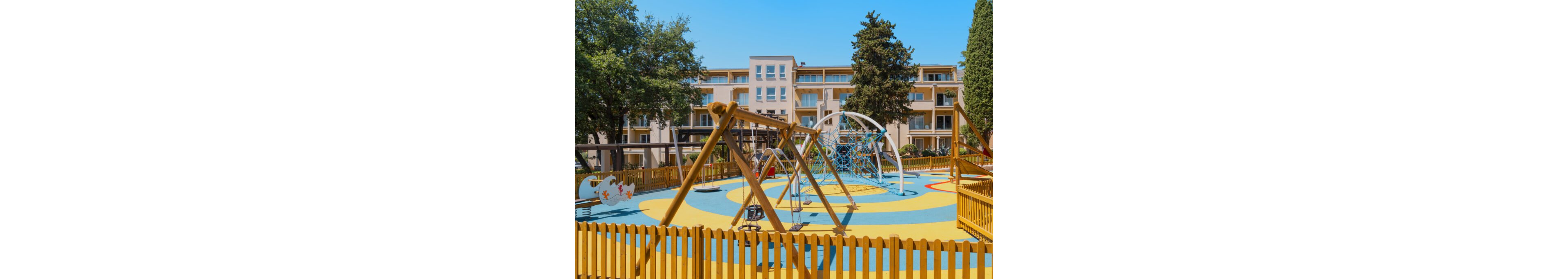 A bright, modern playground is set within a fenced courtyard of a residential apartment complex. The play area features swings, climbing structures, and circular patterns in blue and yellow on the ground. Multi-story buildings with balconies surround the playground, and tall trees provide greenery around the space. The scene is captured on a clear, sunny day, emphasizing a family-friendly urban environment.