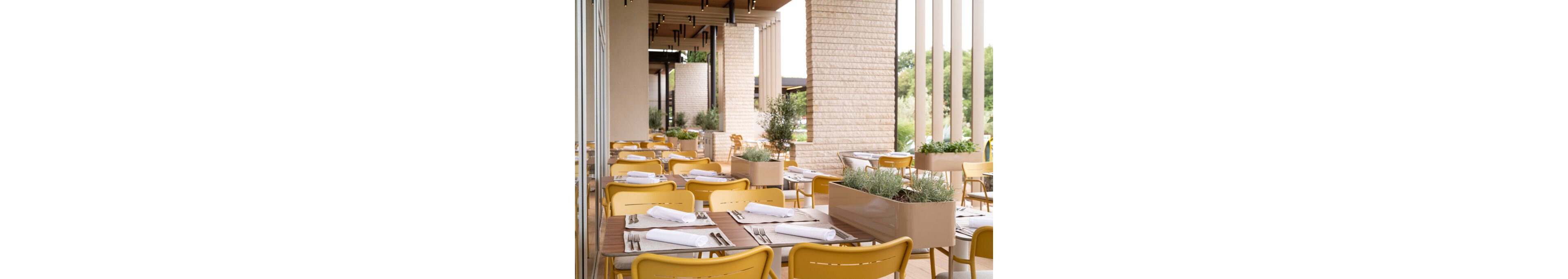 Contemporary outdoor dining area with neatly arranged tables and yellow chairs. Each table is set with white napkins and cutlery, ready for guests. Rectangular planters with green plants separate sections of the terrace and add natural accents. Light-colored stone columns and slatted structures frame the space, with greenery visible in the background.