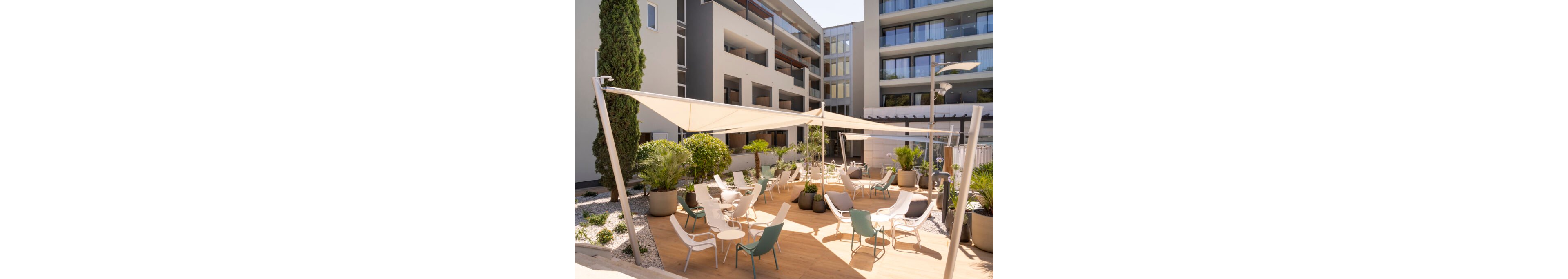Contemporary outdoor seating area is set within a modern apartment or hotel complex. White shade sails stretch above wooden decking, creating a comfortable lounge space with chairs and tables. Large potted plants and gravel beds add greenery and texture to the minimalist architecture. Sunlit balconies and glass railings frame the courtyard, suggesting a relaxed, urban resort atmosphere.