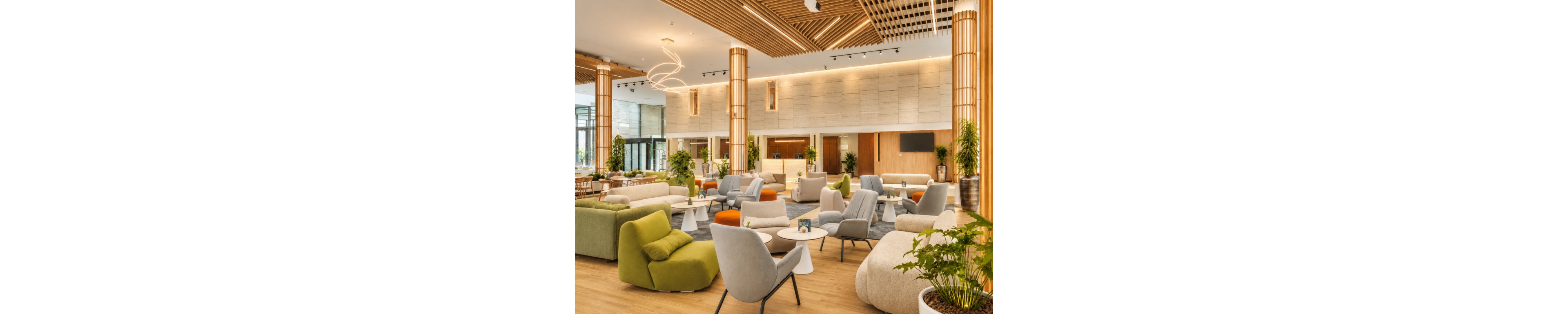 Spacious contemporary lobby interior featuring a variety of modern lounge chairs and sofas in soft neutral and green tones. The setting includes wooden columns, warm ambient lighting, and large windows that bring in natural daylight. A reception desk is visible in the background, complemented by plants and minimalist decor. The overall atmosphere feels welcoming, stylish, and comfortable for guests.