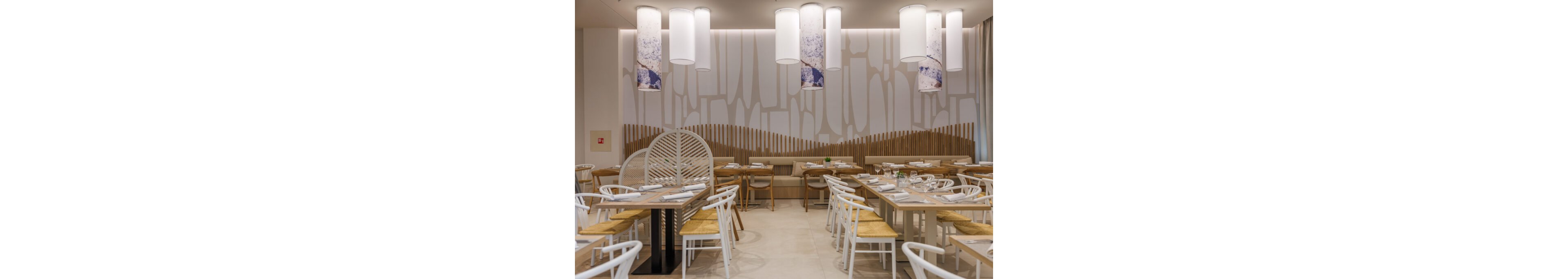 Contemporary restaurant dining area featuring light wooden tables and white chairs with woven seats. The space is bright and airy, with large cylindrical pendant lights hanging from the ceiling. A decorative wall with abstract patterns and vertical wooden slats adds visual interest to the room. Table settings with plates, glasses, and napkins are neatly arranged, creating a clean and inviting atmosphere.
