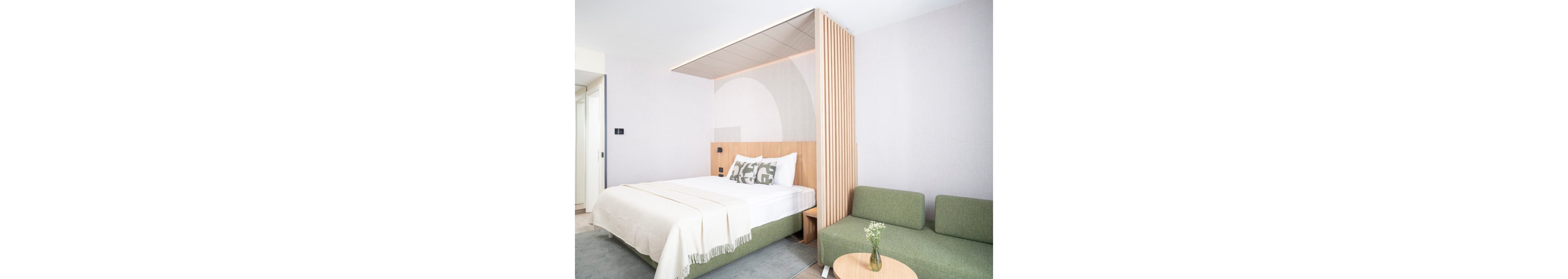 Bright contemporary bedroom features a neatly made double bed with white linens and patterned pillows. A light wood headboard and vertical slatted divider separate the sleeping area from a green upholstered sofa. Soft neutral walls and a pale rug create a clean, minimalist aesthetic. A small round table with a glass vase and flowers adds a subtle decorative touch.