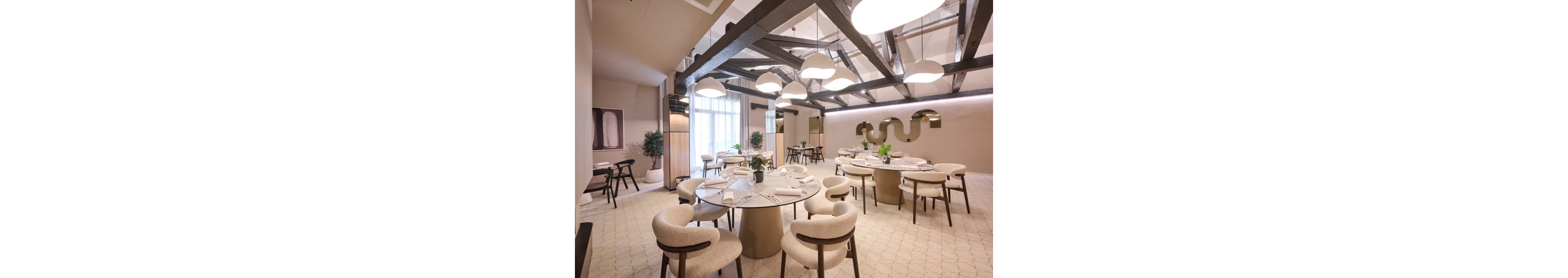 Contemporary restaurant dining room featuring round tables and upholstered chairs under exposed wooden ceiling beams. Soft pendant lighting creates a warm, inviting atmosphere with neutral tones and minimalist decor. Large windows with sheer curtains bring in natural light, while plants and abstract wall art add visual interest. The space appears prepared for guests, with neatly arranged table settings and a clean, spacious layout.