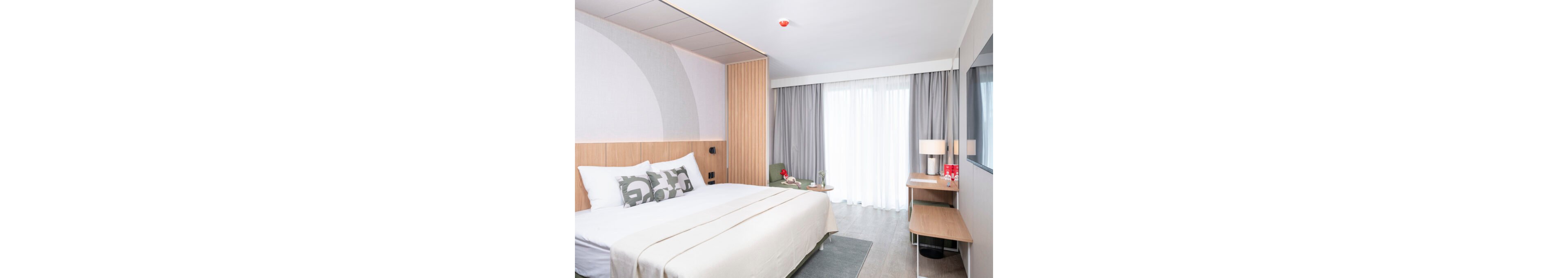 A contemporary hotel room features a large neatly made bed with white linens and patterned pillows. Light wood paneling and soft gray curtains frame a wide window with sheer drapes, filling the space with natural light. A minimalist desk with a lamp, chair, and small decorative items sits against the wall opposite the bed. The overall design is clean and modern, with neutral tones and subtle accents creating a calm atmosphere.