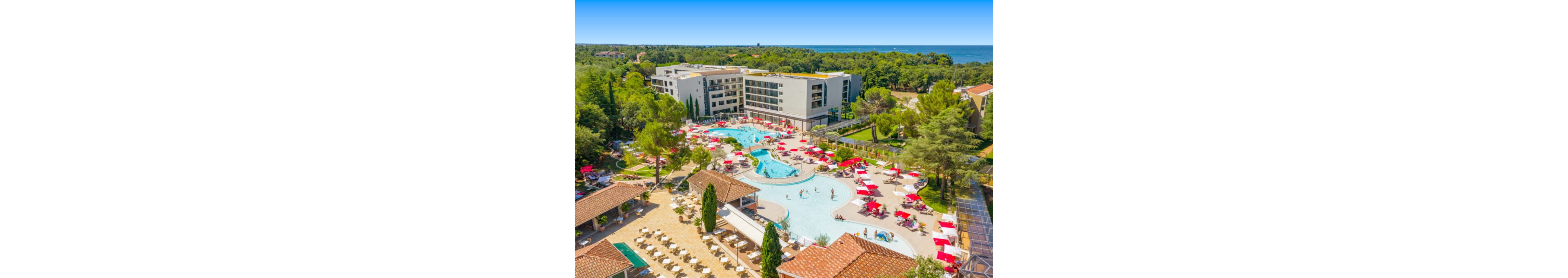 A large holiday resort features a central outdoor pool complex surrounded by modern hotel buildings and lush greenery. Guests relax on sun loungers under red and white umbrellas and enjoy swimming in the curved, light-blue pools. The scene includes multiple terraces, pathways, and landscaped trees, with a forested area and sea visible in the distance. Bright daylight and clear blue sky create a vibrant, summery atmosphere.