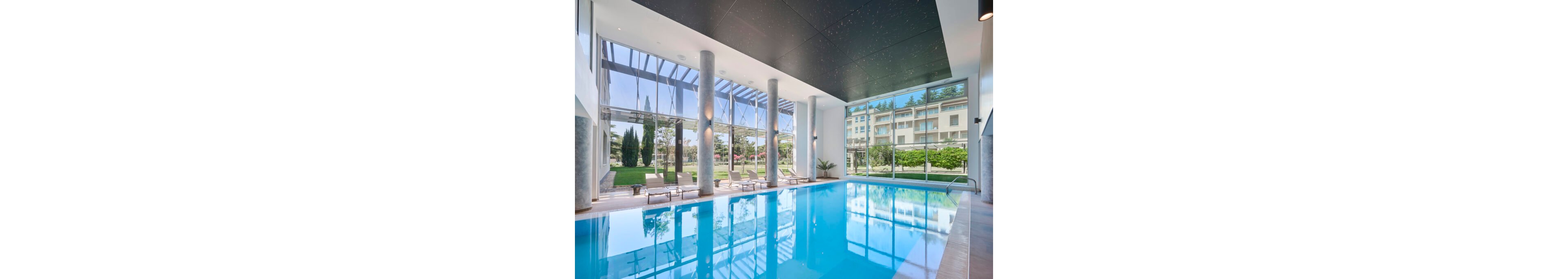 A bright indoor swimming pool is set within a contemporary building featuring tall glass windows and concrete columns. Sun loungers line the poolside, facing out toward a landscaped garden with trees and manicured lawns. Natural light floods the space through the expansive glazing and overhead structure, creating reflections on the calm blue water. In the background, a multi-story residential or hotel building is visible beyond the garden.