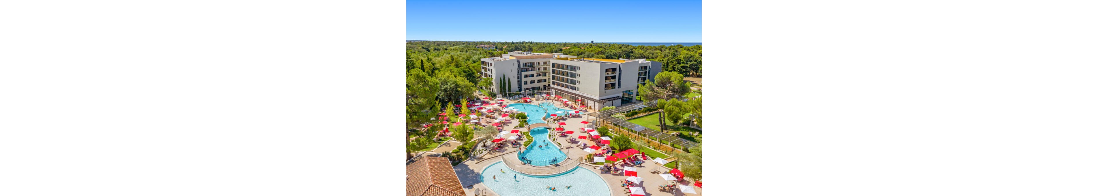 A modern resort hotel features multiple outdoor swimming pools surrounded by sun loungers and umbrellas. Guests relax and play in the water under clear blue skies, with landscaped gardens and trees framing the property. The building has several floors with balconies overlooking the pool area and nearby recreational spaces. In the distance, dense greenery and a hint of coastline complete the vacation setting.