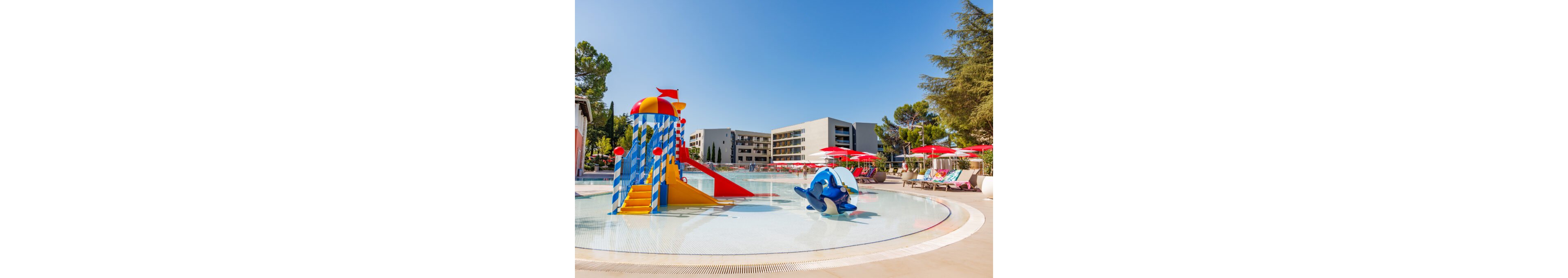 An outdoor splash pool at a resort features colorful water slides and playful structures under a clear blue sky. A striped slide tower with bright red and yellow accents stands in shallow water, alongside a blue dolphin-shaped play element. Red umbrellas shade lounge chairs around the pool, with modern apartment-style buildings visible in the background. Tall trees frame the scene, creating a relaxed vacation atmosphere.