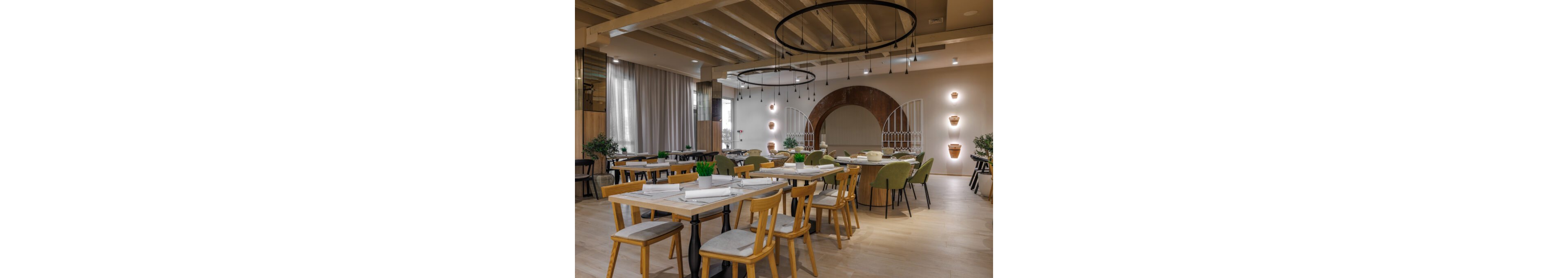 Contemporary restaurant dining area featuring wooden tables and mixed seating with green and beige upholstered chairs. The space includes decorative wall arches, potted plants, and soft wall lighting that creates a warm ambiance. Circular pendant lights hang from the ceiling beams, illuminating neatly arranged place settings on the tables. Neutral tones and clean lines give the interior a stylish yet inviting atmosphere suitable for casual or fine dining.