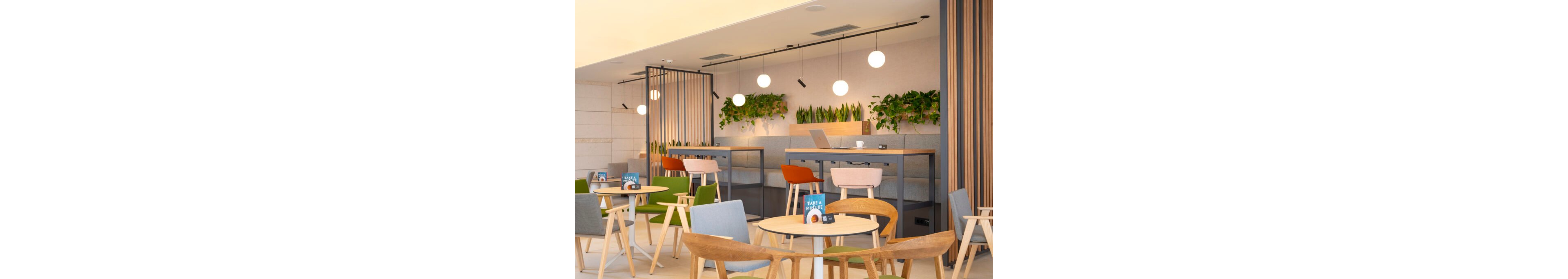 Contemporary cafe interior featuring light wooden tables and colorful chairs. High counter seating with laptops suggests a casual workspace environment. Green plants along the walls and hanging planters add a natural touch to the bright space. Warm lighting from round pendant lamps creates a welcoming and relaxed atmosphere.