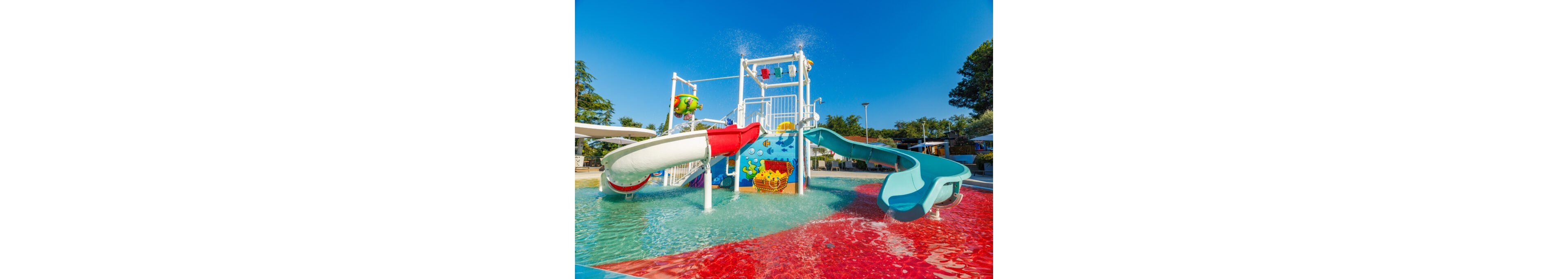 A vibrant outdoor water playground features two curved slides emptying into a shallow splash pool. The central structure includes water buckets and sprayers, decorated with playful cartoon sea creatures. Bright red and blue pool surfaces contrast with the clear turquoise water under a sunny blue sky. Trees and resort-style seating are visible in the background, suggesting a family-friendly recreational area.