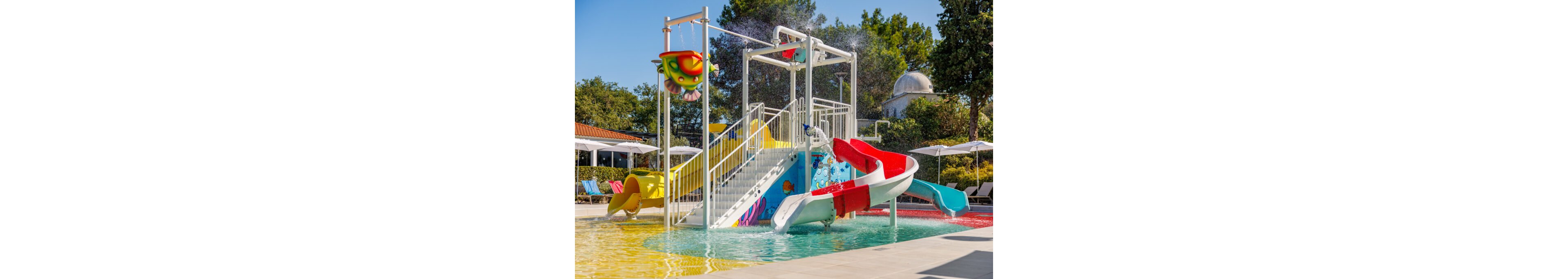 A vibrant outdoor water playground features multiple slides and splash structures in a shallow pool. A red and white curved slide and a yellow slide descend from a white metal platform with stairs and railings. Colorful water buckets and playful graphics decorate the central structure, while water sprays create a fun, refreshing atmosphere. Sun loungers and umbrellas line the background, surrounded by trees and a bright blue sky.