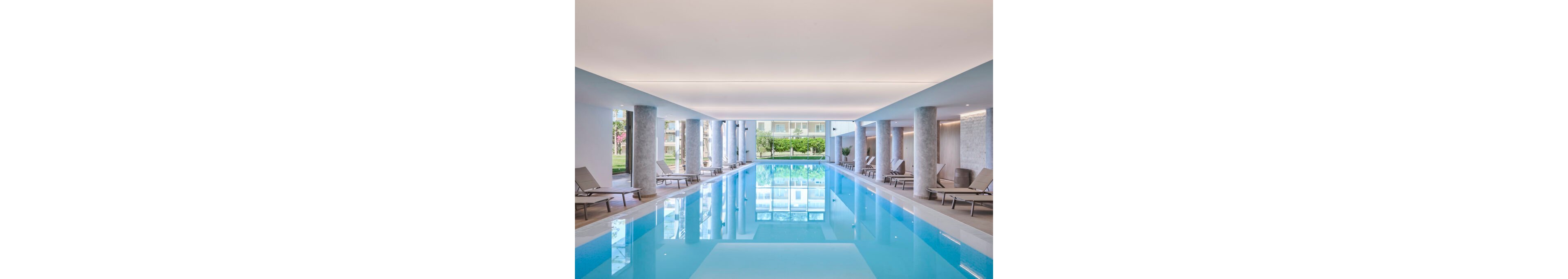 A long, rectangular indoor swimming pool stretches through a bright, contemporary space. Concrete columns line both sides, with beige lounge chairs neatly arranged along the pool deck. Large openings at the far end reveal greenery and residential buildings outside, bringing natural light into the area. The clean design and soft lighting create a calm, relaxing atmosphere suitable for leisure and wellness.