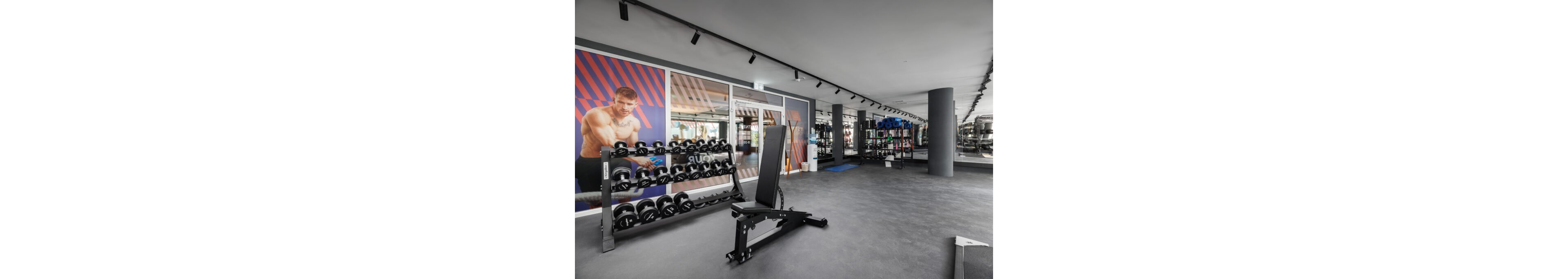 A spacious, contemporary gym features a bench and rows of dumbbells in the foreground. Large windows and mirrors line the wall, reflecting additional fitness equipment and storage racks. A colorful wall graphic of a muscular man exercising adds visual interest to the space. The clean, minimalist design and neutral tones create an open, professional workout environment.
