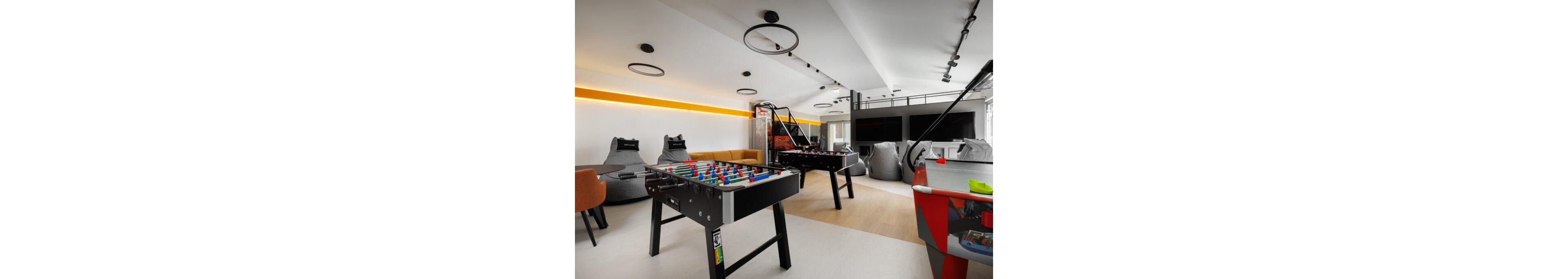 A contemporary office recreation room features multiple game tables, including foosball and air hockey, arranged in an open, bright space. Sleek seating options such as bean bags, lounge chairs, and a sectional sofa provide relaxed areas for employees to unwind. The room is accented by minimalist circular ceiling lights and a horizontal strip of warm yellow lighting along the wall. Large screens and gaming machines in the background enhance the entertainment-focused atmosphere.