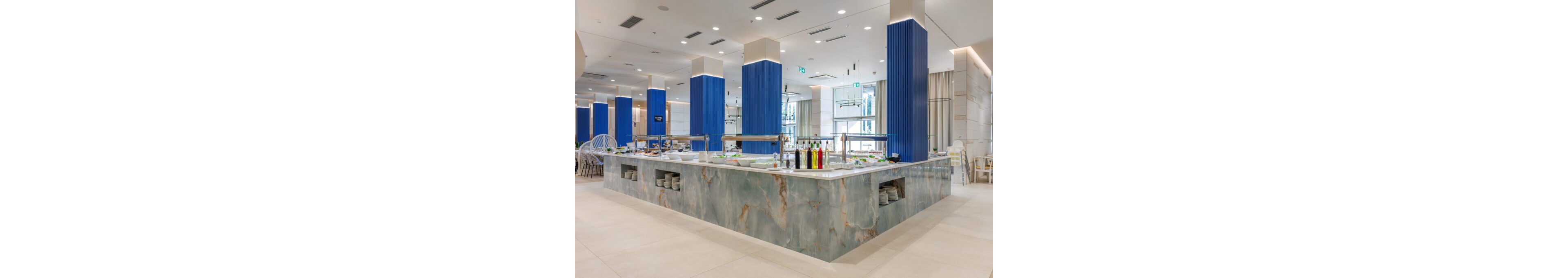 A large marble-style buffet counter stands in the center of a bright, modern restaurant interior. Blue vertical columns and light tiled floors create a clean, contemporary atmosphere. Serving stations with trays, plates, and condiment bottles line the countertop, with shelves of dishes visible underneath. The space is well-lit with recessed ceiling lights and surrounded by dining tables and chairs.