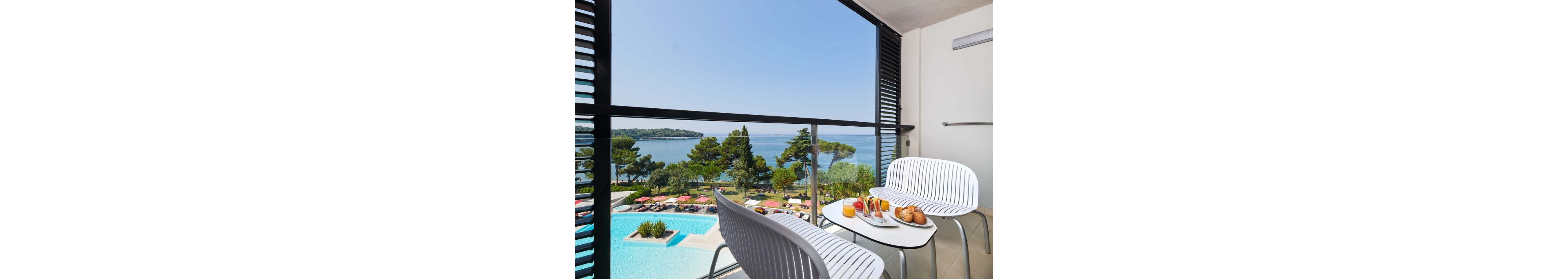 A modern balcony with two white chairs and a small table overlooks a coastal resort. On the table are glasses of juice, coffee, and a plate of pastries set for breakfast. Below, a large swimming pool, sun loungers, and umbrellas sit among landscaped greenery. Beyond the resort, calm blue sea and a tree-covered island stretch into the distance under a clear sky.